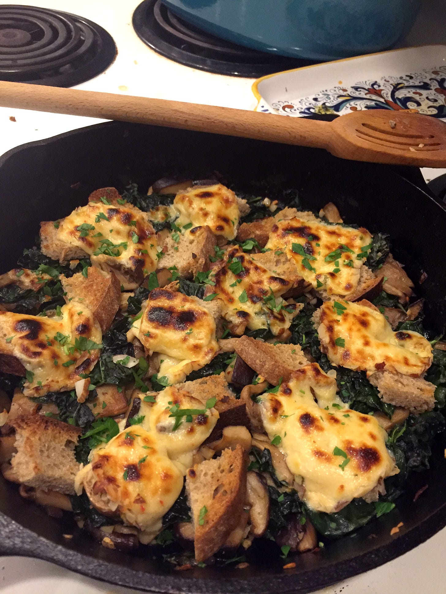 a cast iron pan full of croutons, kale, and mushrooms, with browned slices of pepper jack cheese on top. A slotted wooden spoon sits on the edge of the pan.