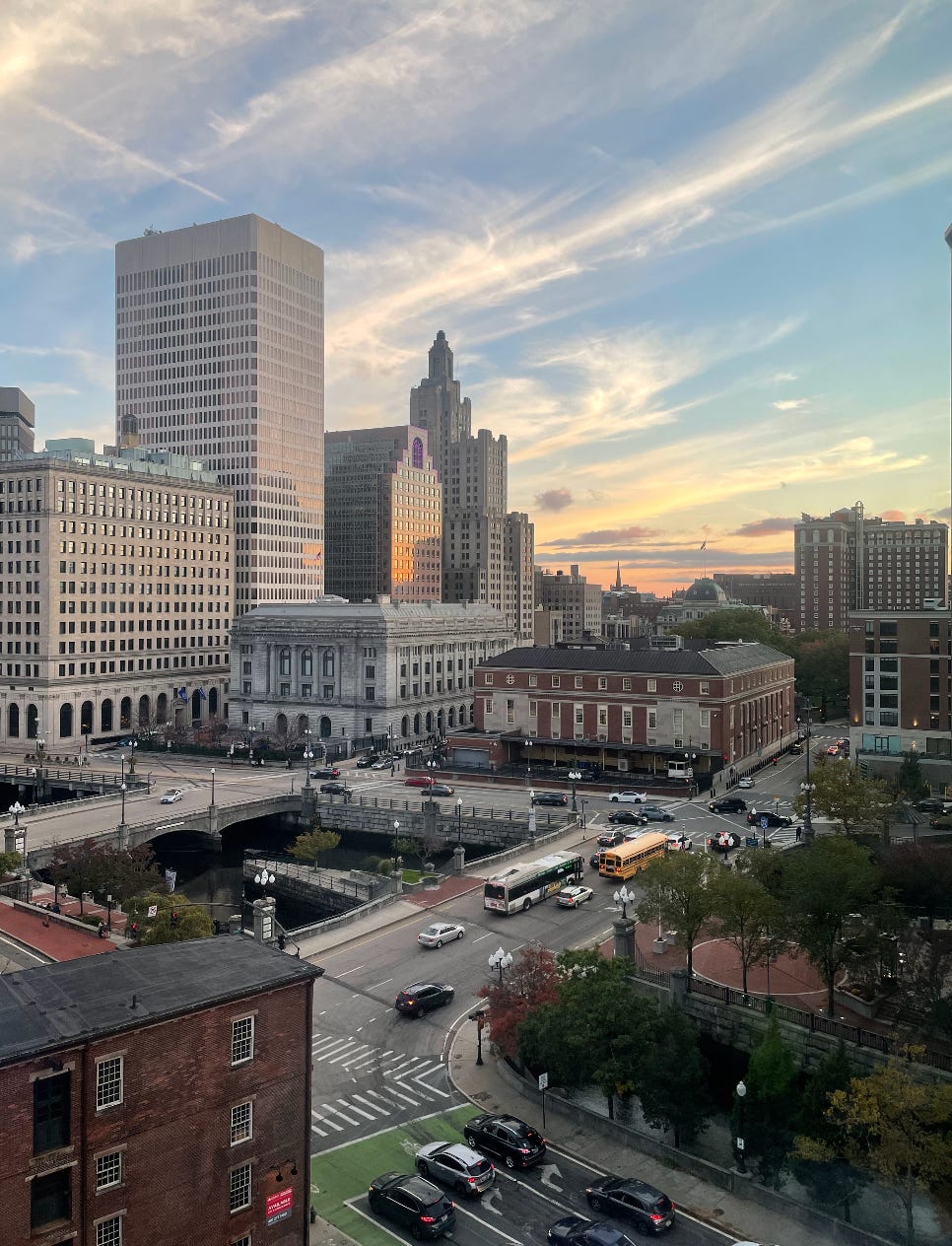 The providence skyline during a golden hour sunset. The sky is streaked with lies of clouds and purple, blue and yellow hues.