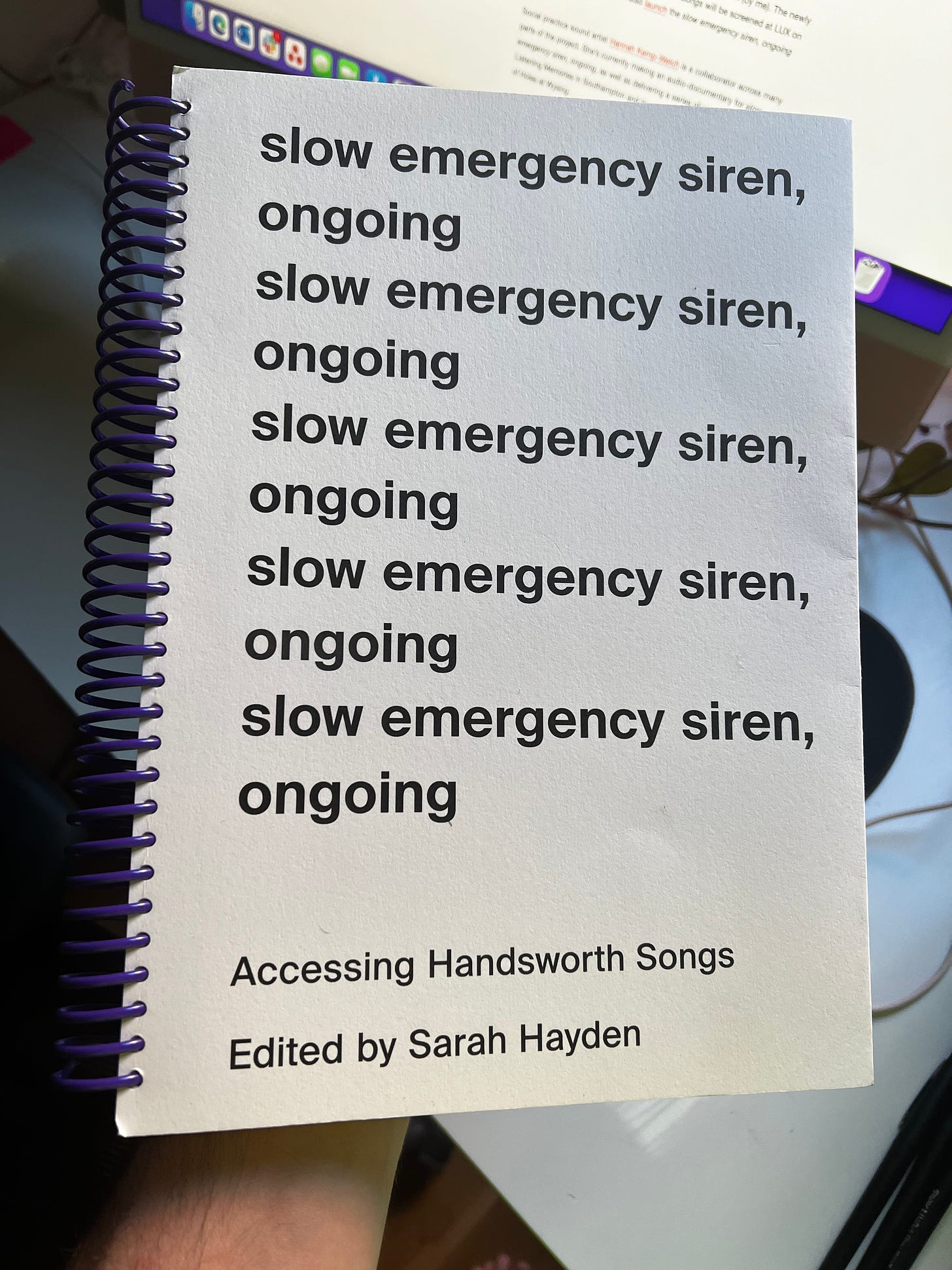 I’m holding a book with a purple spiral binding. Black bold, lower-case text repeats the title: slow emergency siren, ongoing. At the bottom, smaller: Accessing Handsworth Songs / Edited by Sarah Hayden.