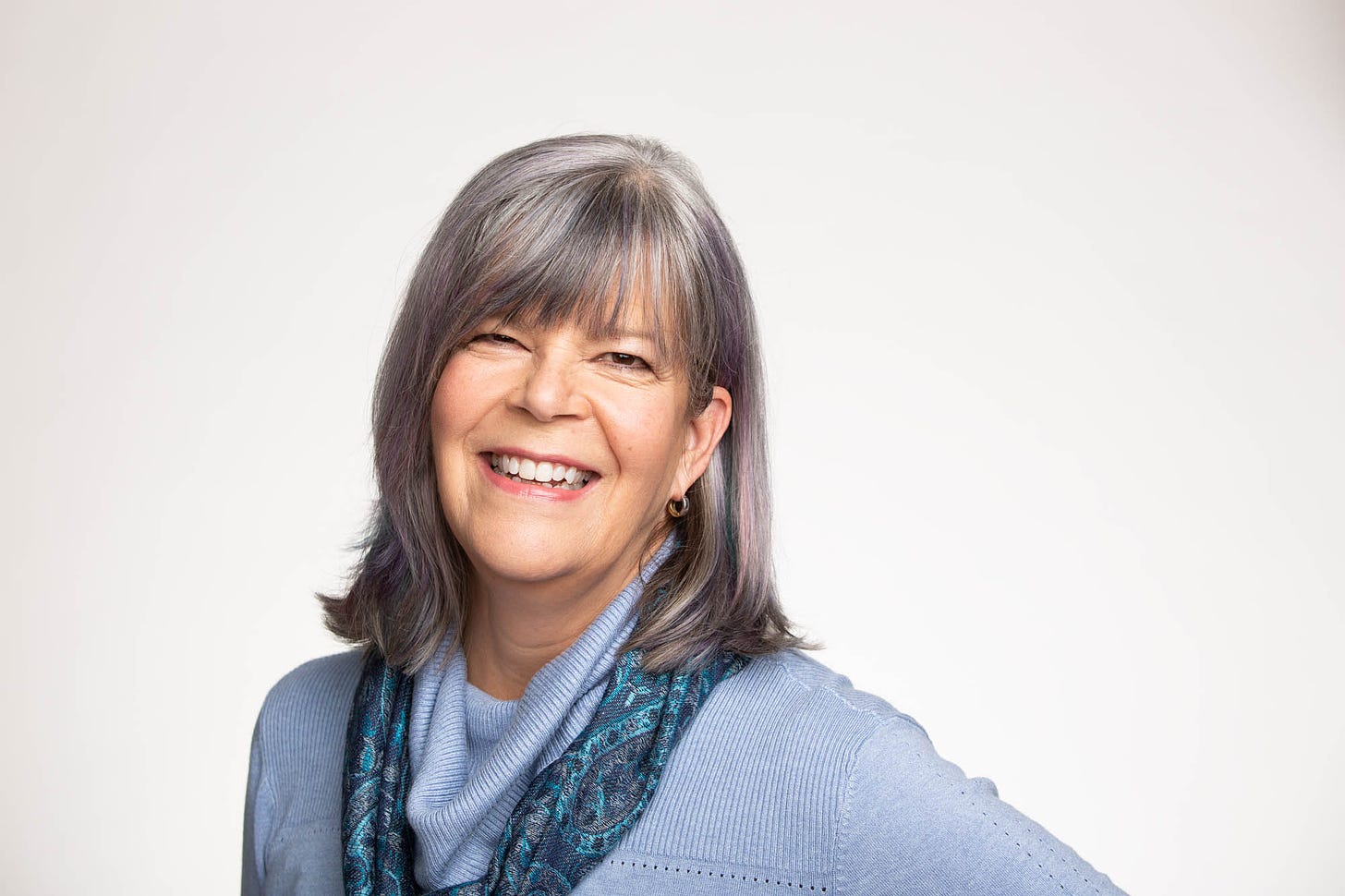 Karen is a white woman with shoulder-length silver hair with a fringe. She's wearing a pale blue jumper with a patterned blue scarf and smiling at the camera against a plain white background.