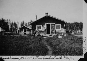 Photo caption: "Sizeland's Roadhouse, Nenana - Fairbanks [Trail? Train?], 6/20/21 [6/20/22]" Surrounding photographs in this collection are dated in year, 1922, so 1921 may be incorrect. This photo shows a man standing with his hands on his hips by the doorway of a cabin facing two other men sitting down. There's a chair to the right of the doorway. Two smaller cabins are located to the left. A dirt path leading up to the larger cabin has been worn away through a dense patch of brush. [UAF-1969-89-108]