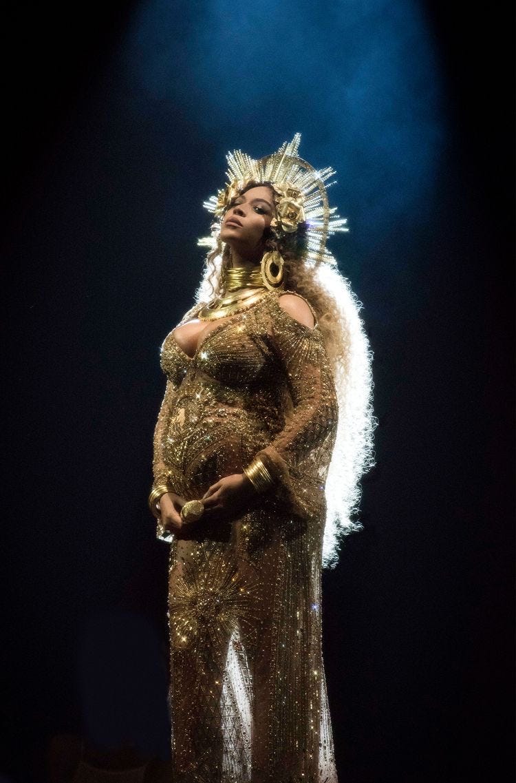 Beyonce, crowned and golden, at the 2017 Grammys. She wears a golden radial crown, stacked thick golden jewelry, and a golden mesh dress falling just right over her (very pregnant with twins) figure.
