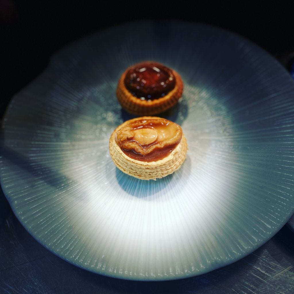 A sideview of a cooked abalone encase in a pastry tart case in the centre of a textured dinner plate