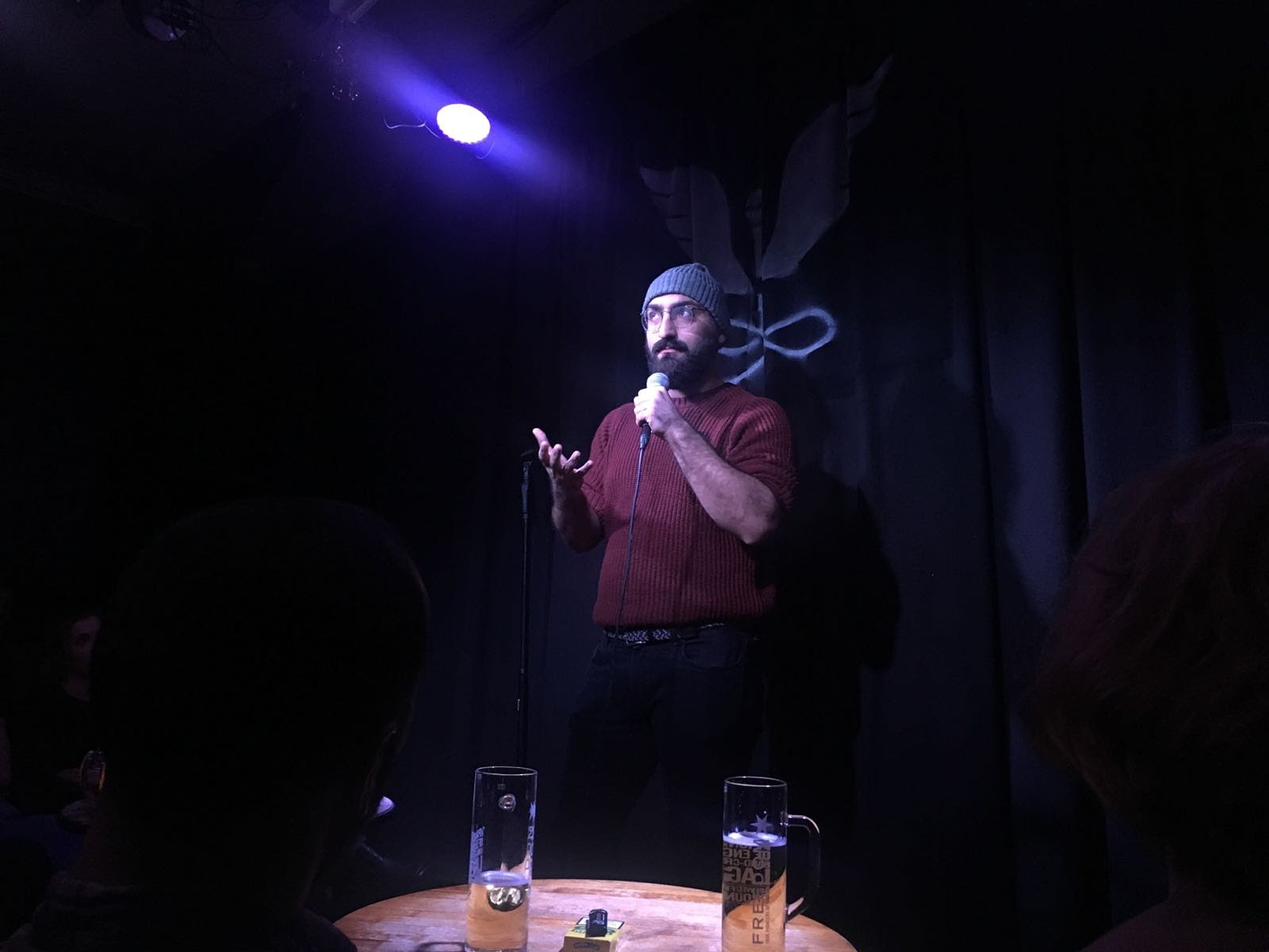 Amir on stage in an orange jumper and grey hat holding a mic in front of the Angel Comedy logo. Two half finished pints from the audience are visible on a table in front of the stage.