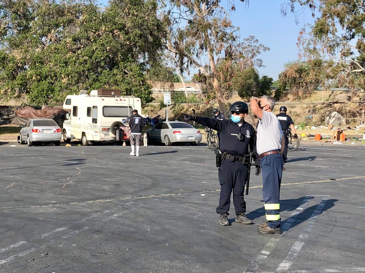 A police officer in the foreground, in the background several broken down vehicles and a homeless encampment.