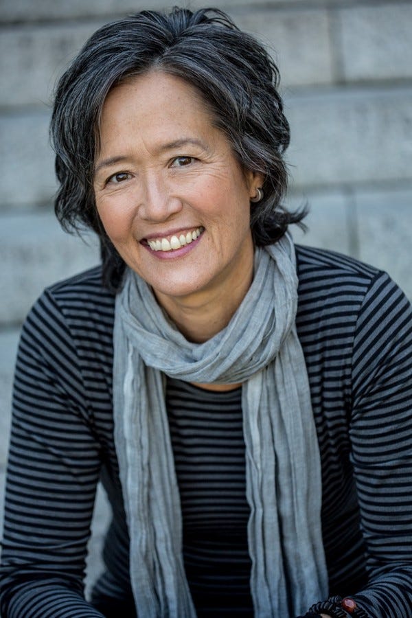 Portrait shot of author Ruth Ozeki wearing a grey scarf and black and grey striped shirt, smiling at the camera