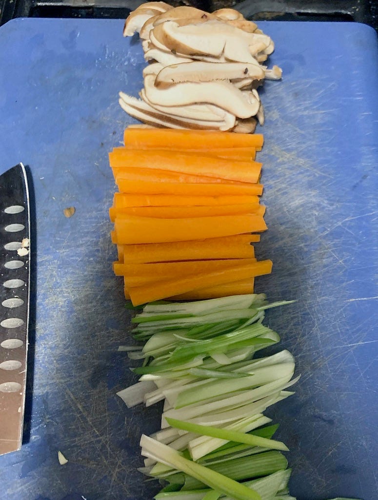 Image shows a blue cutting board arranged with a line of julienned scallions, carrots, and sliced shiitake mushrooms arranged down the center. To the left side, there is a santoku-style knife resting.
