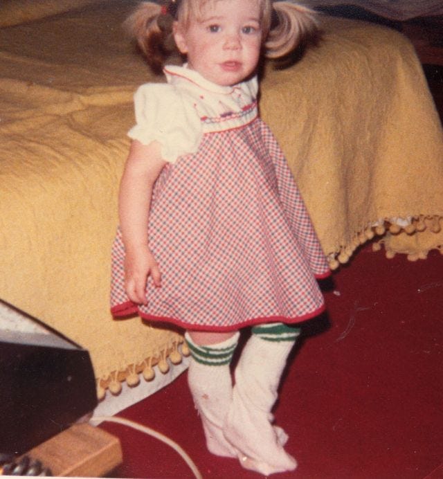 Young Emily stands next to a bed wearing a dress and a pair of too-large athletic socks. Her hair is in pigtails and her face is dirty