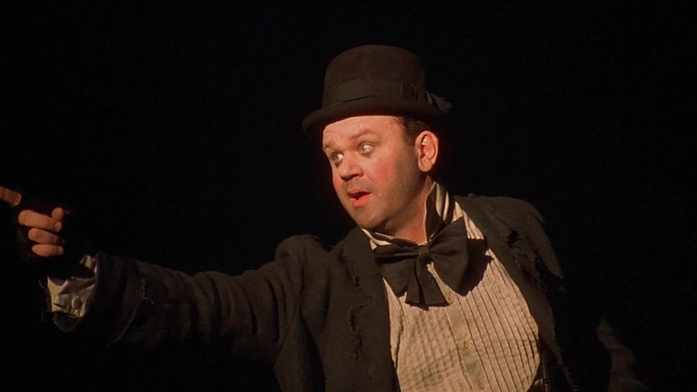 John C. Reilly wears a bowler hat, ragged coat, large bow tie and fingerless gloves and is pointing and looking off to the side.