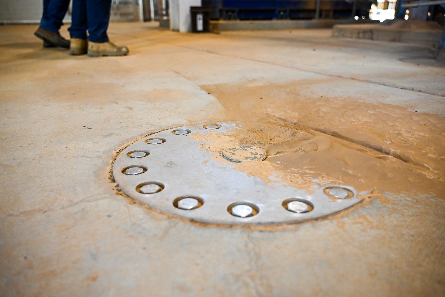 a close up of a concrete floor. in the foreground is a bolted silver cover, unknown what lies beneath, but a beige mix of sand and ash accumulate on the right edge of the cover where the bolts meet the concrete. two employees' work boots are blurred in the distance.