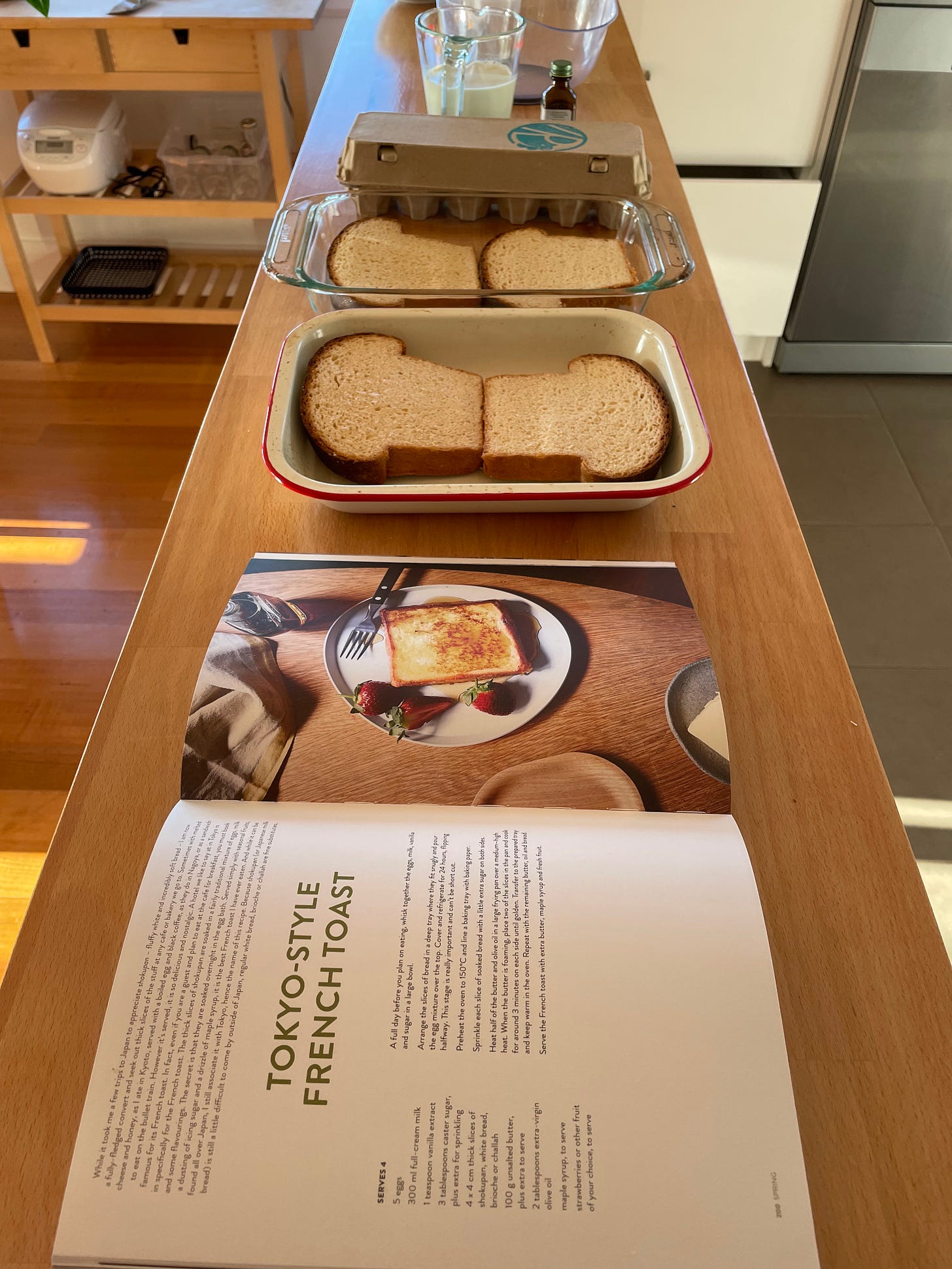 Two trays of milk bread on a kitchen counter with a cookbook open at Tokyo French Toast. Milk, eggs and vanilla essence in the background.