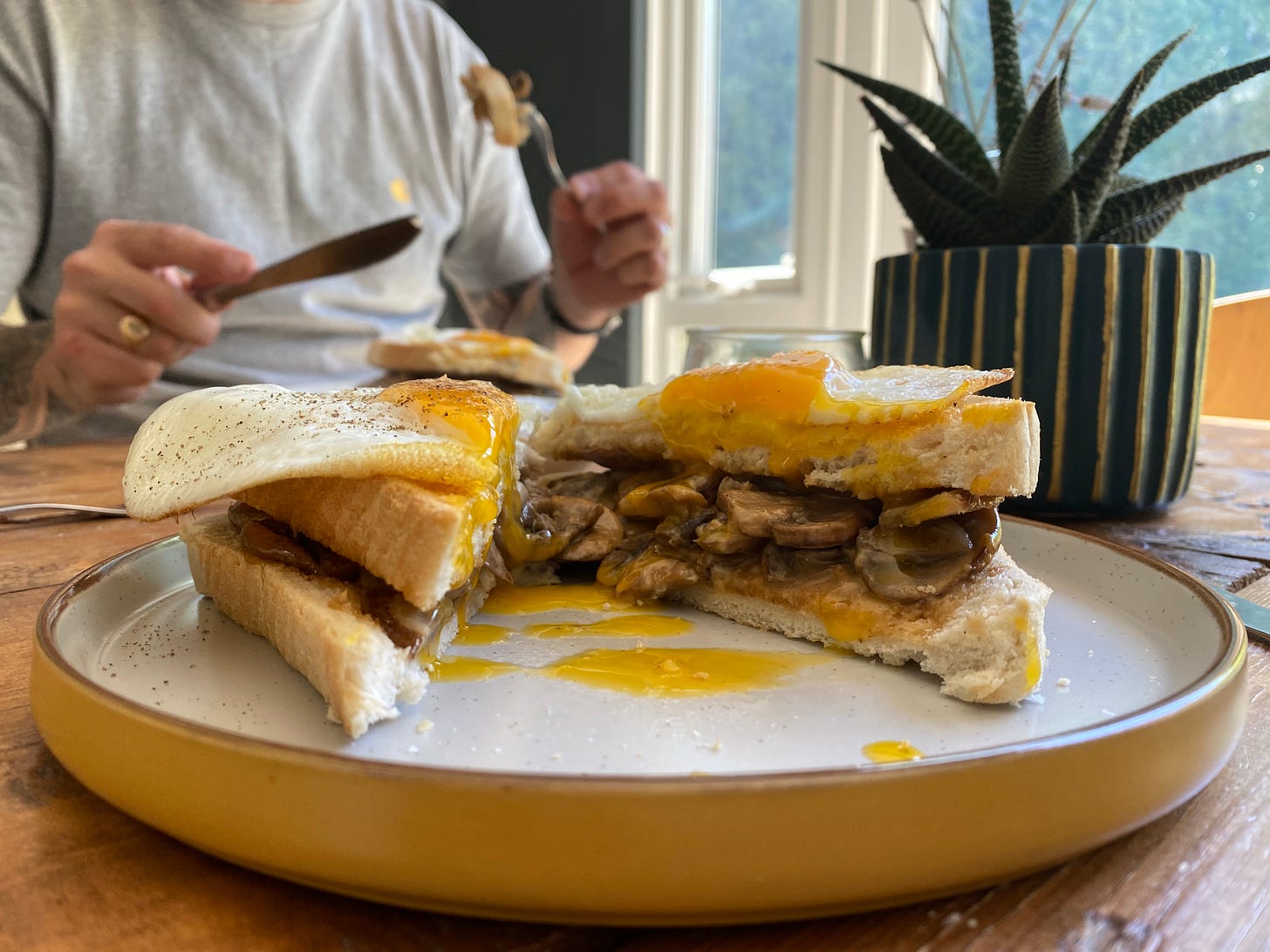 Plate on a table with a cut open sandwich filled with sliced mushrooms. A runny yolk egg is on top, and yellow yolk is splodged over the plate. A person is in the background holding a knife and fork poised to eat.