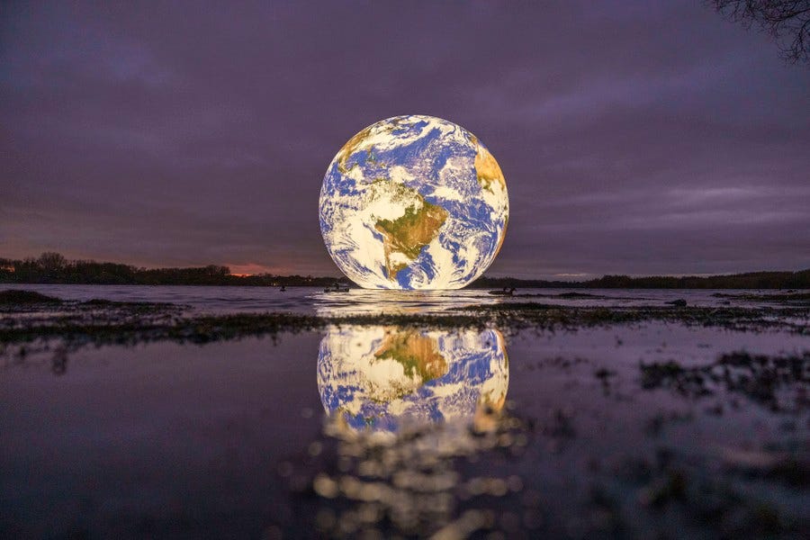 A large inflatable model of the Earth, lit from within, floats in a small lake at sunset.