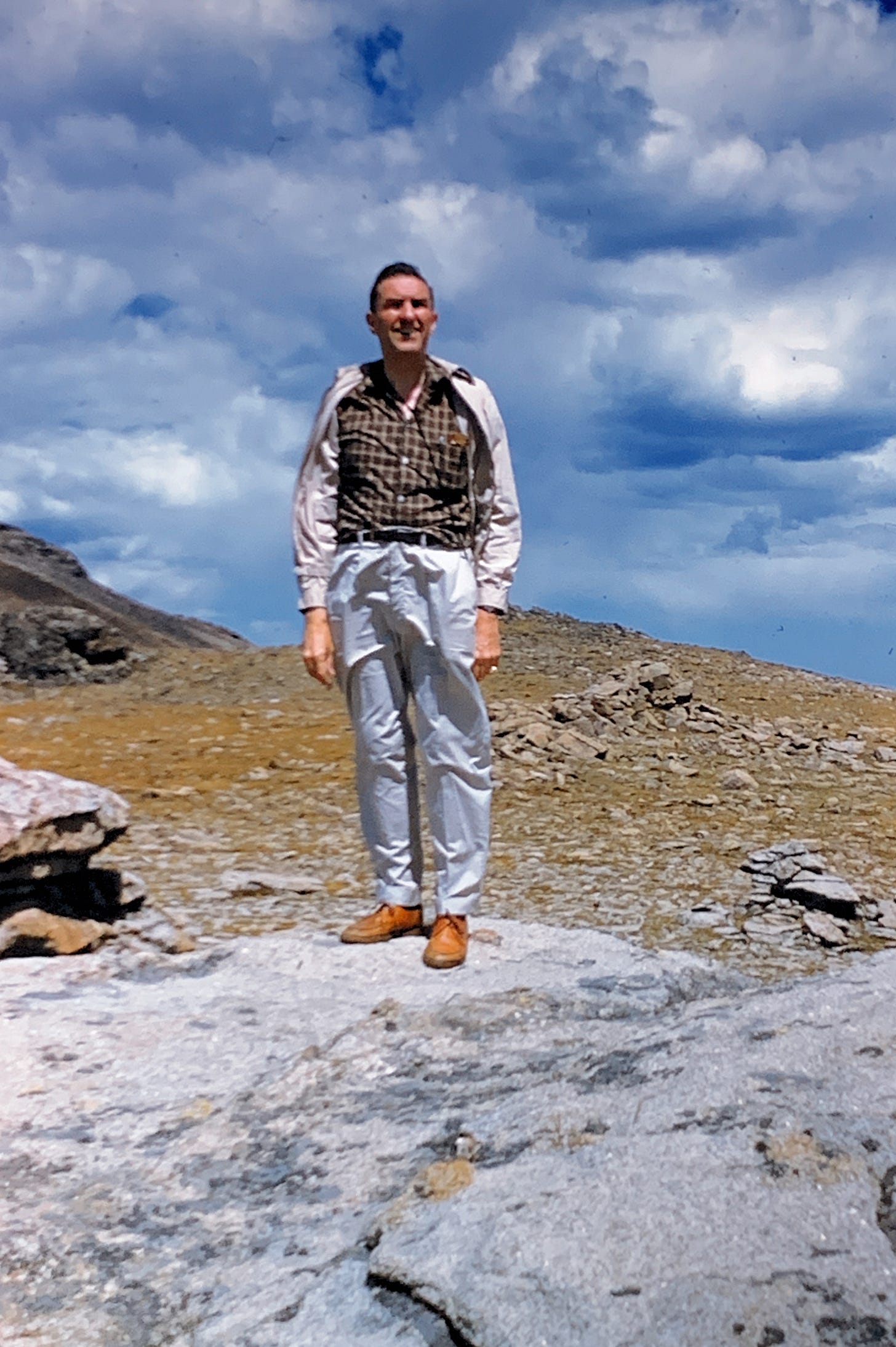 A man standing on a rock on a small hill with larger rocky hills in the background. He is looking towards the camera but above it, with his hands at his sides and a smile on his face.