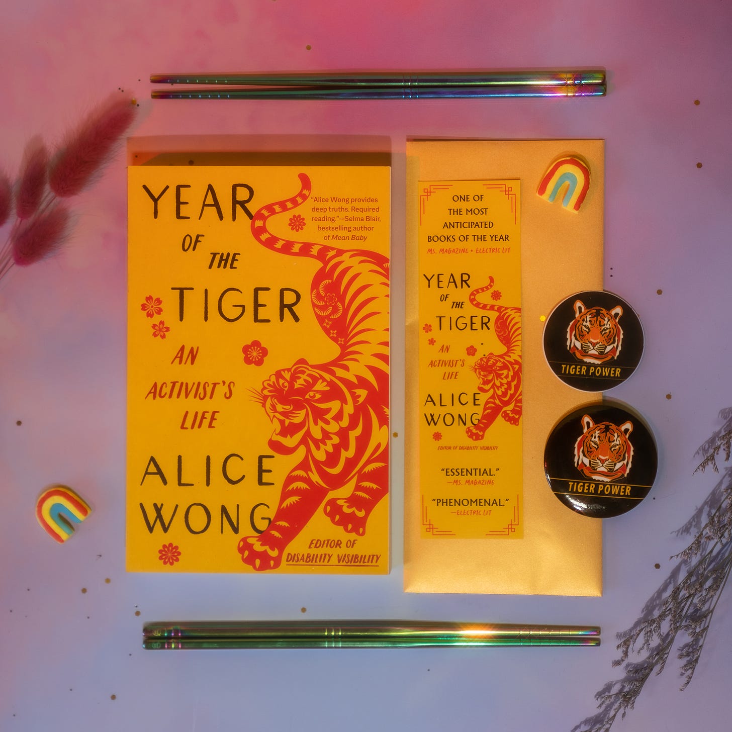 Photo with a purple-pink background with a copy of Year of the Tiger paperback in the center. Above and below the book are a pair of rainbow chopsticks from @umeshiso_ (on IG). On the right of the book is a tiger bookmark and is one round tiger sticker and one round tiger button. Floral decorations and a small rainbow decor are placed around the edges. Photo credit: @ziru.mo (on IG)