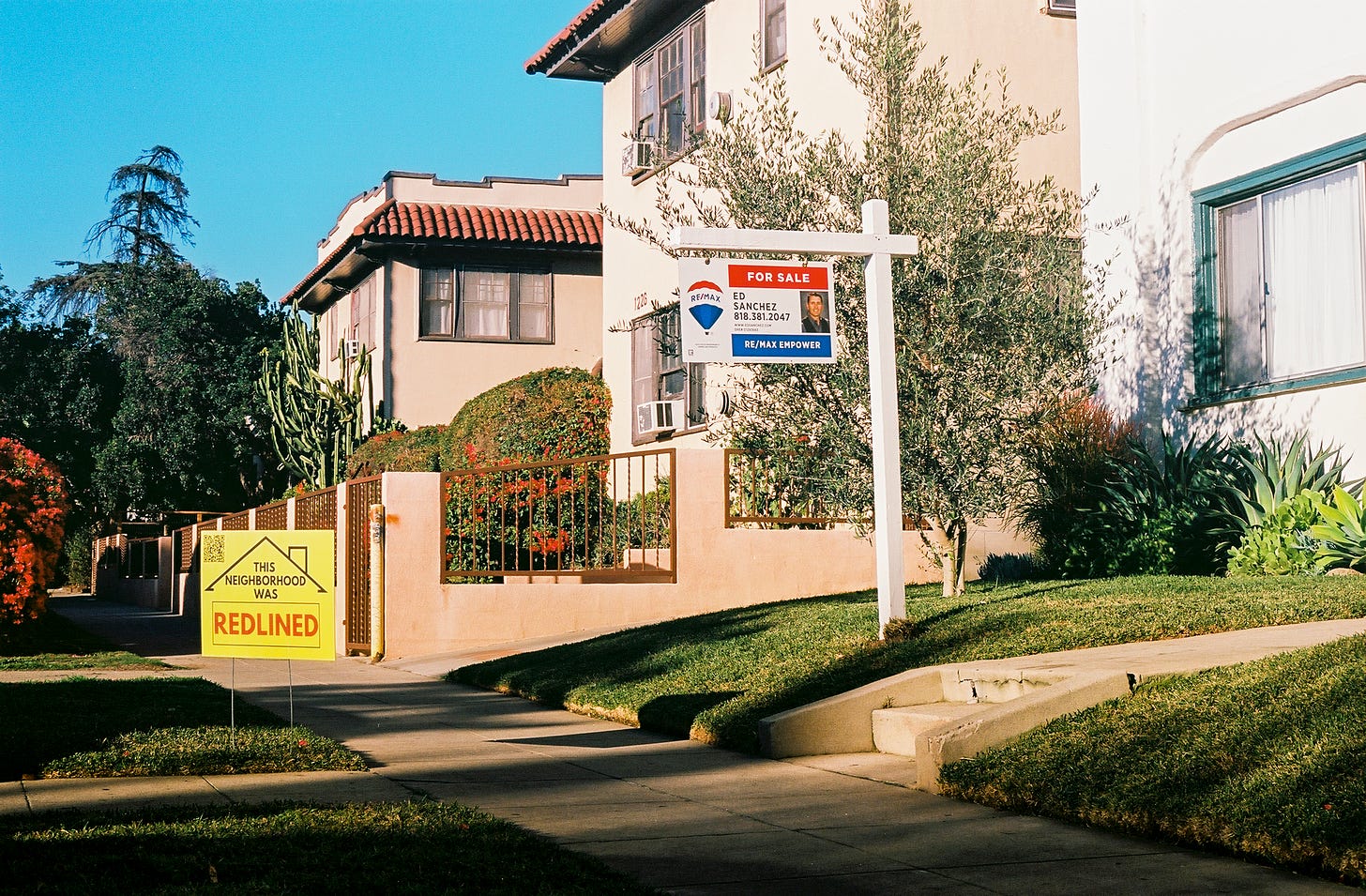 Photo of a yellow redlining yard sign planted in the grass in front of an apartment building that has a for sale sign on it.