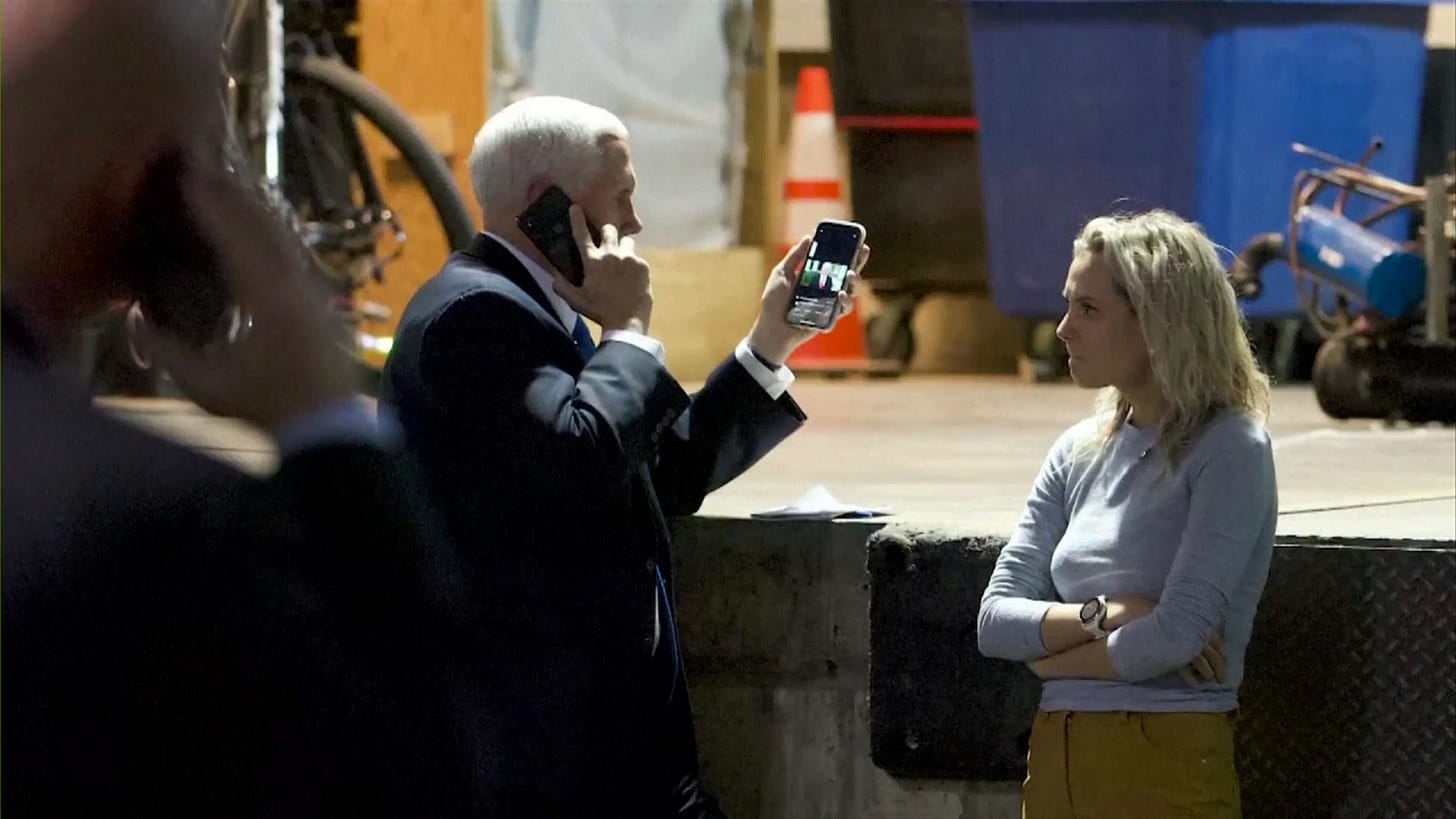 Mike Pence, in the underground secure location, on the phone in his right hand as he watches Trump's video message to his insurrectionists in his left hand. A blonde woman crossed arms looks at him from the right. What appears to be an air compressor or perhaps a welding rig? is in the far background.