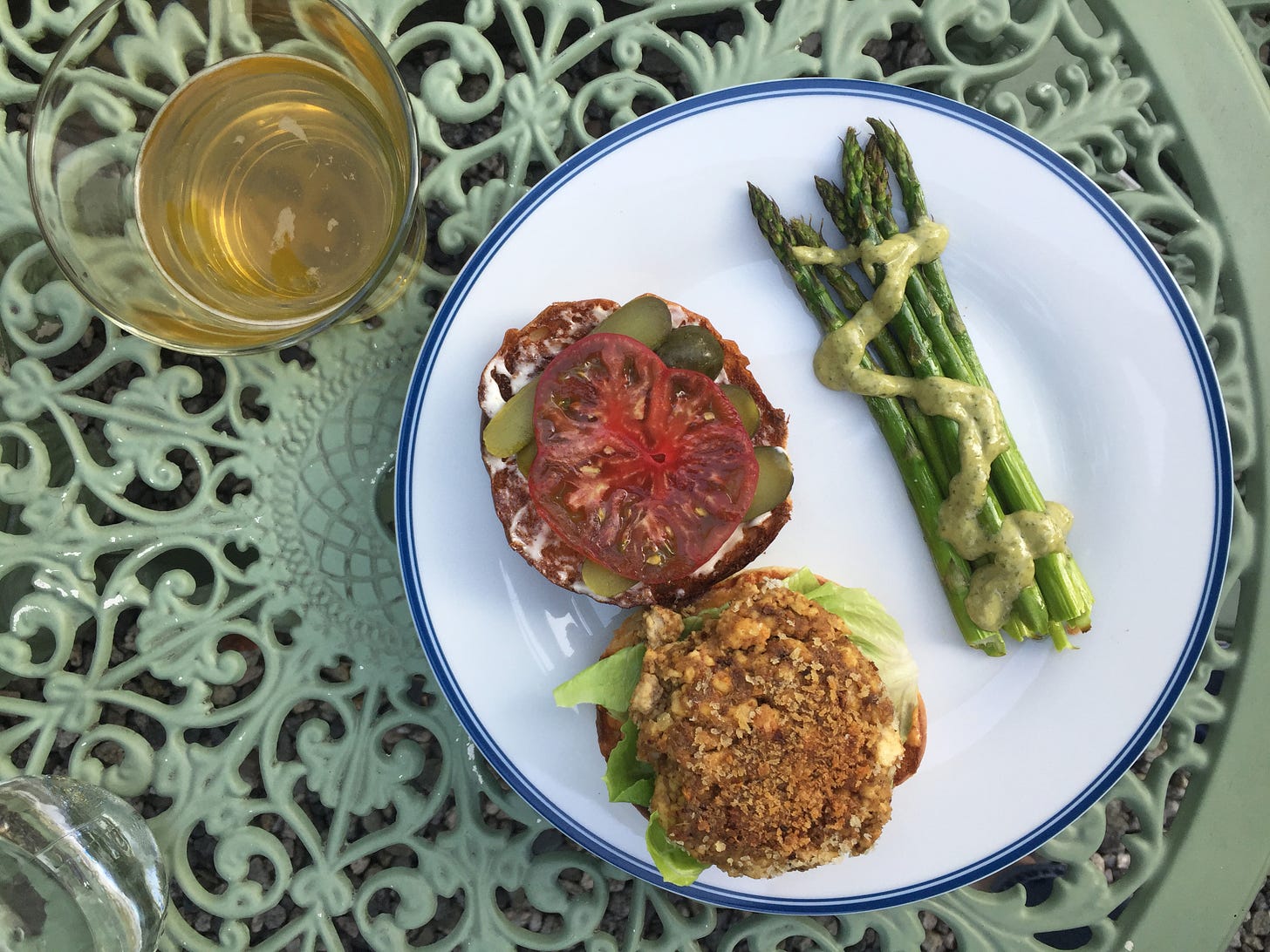 from above, on a green wrought-iron table, two halves of a burger rest on a plate next to spears of asparagus with a drizzle of green dressing. On one half of the burger is the panko-crusted patty on top of lettuce, and on the other half is a slice of heirloom tomato on top of slices of pickles.