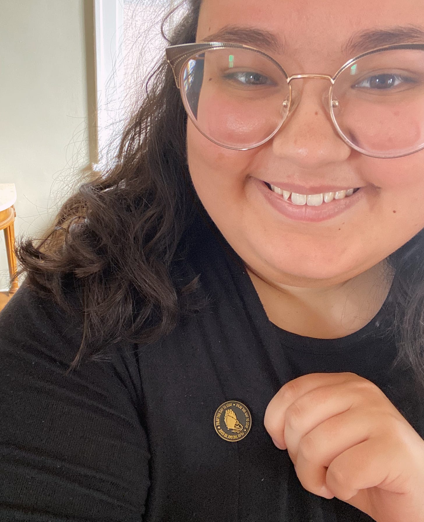  Chloe is smiling into the camera in this close-up shot where she is showing off her lapel pin which is black with gold accents. The lapel pin features hands folded in prayer and holding rosary beads in the center of the design. Around the hands are the words “pray the gay to stay”, which is printed 3 times around the edges of the pin. 