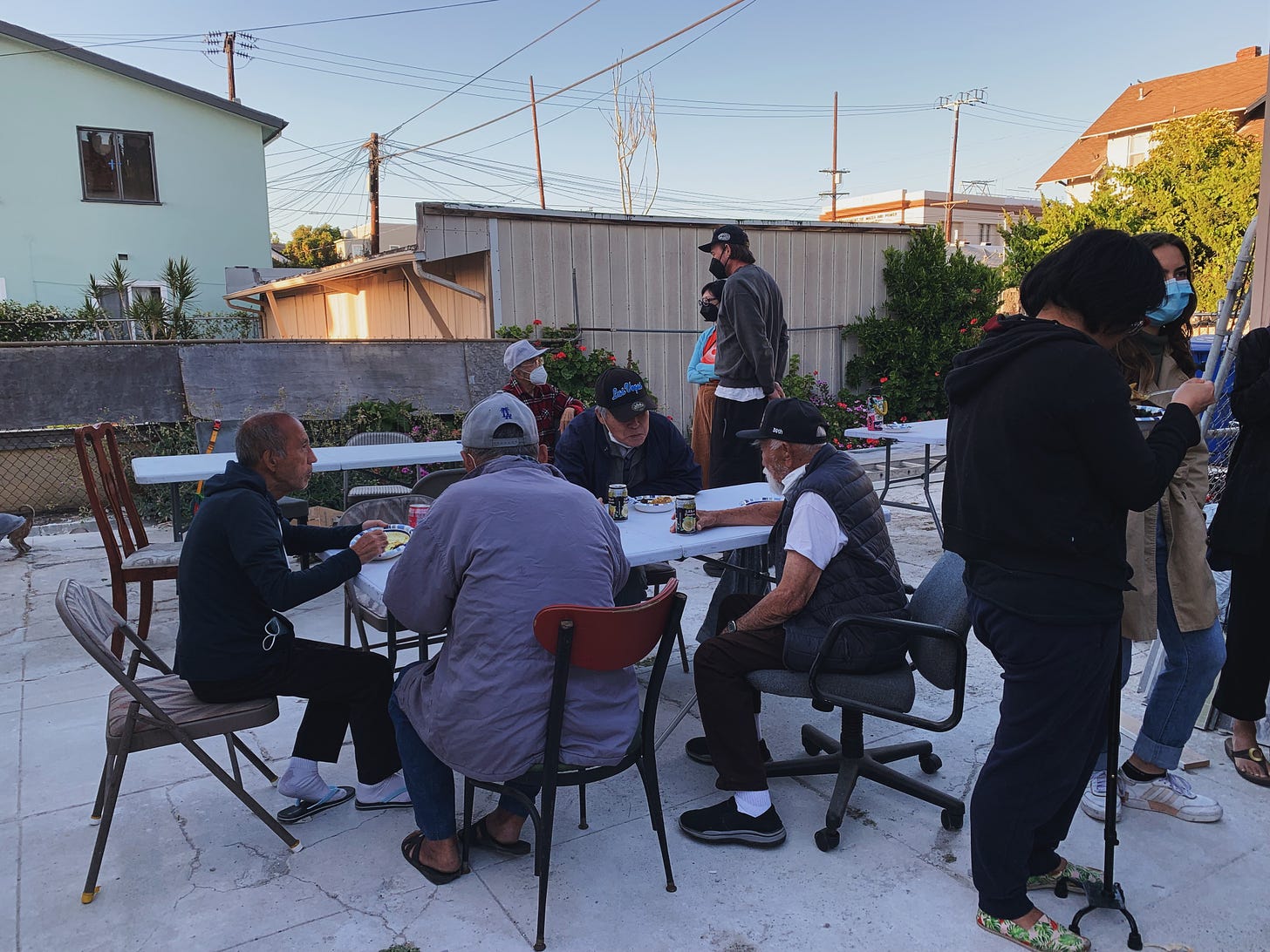 four elder Japanese tenants seated at a table and having a meal outside on a slab of concrete, surrounded by organizers and community members with apartments and electric poles in the background 