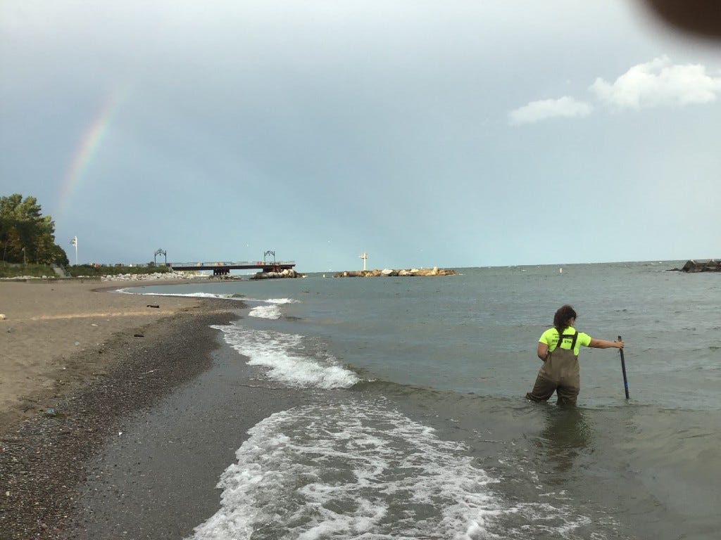 A woman in a Sewer District uniform stands knee deep in Lake Erie as she holds a sample stick stabilizing her for safety. She faces away from the camera as in the distance a rainbow peeks through the clouds after a morning storm.