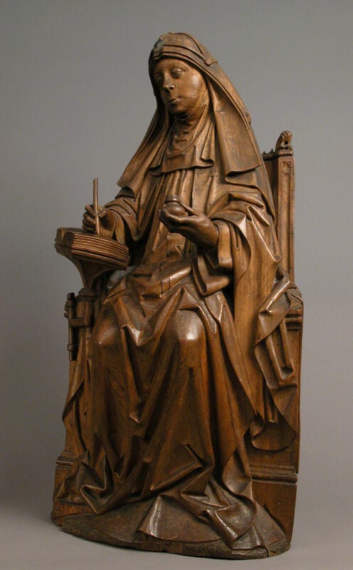 En träskulptur av en kvinna med dok. Hon sitter och skriver med ett bläckhorn i ena handen och penna i den andra.
