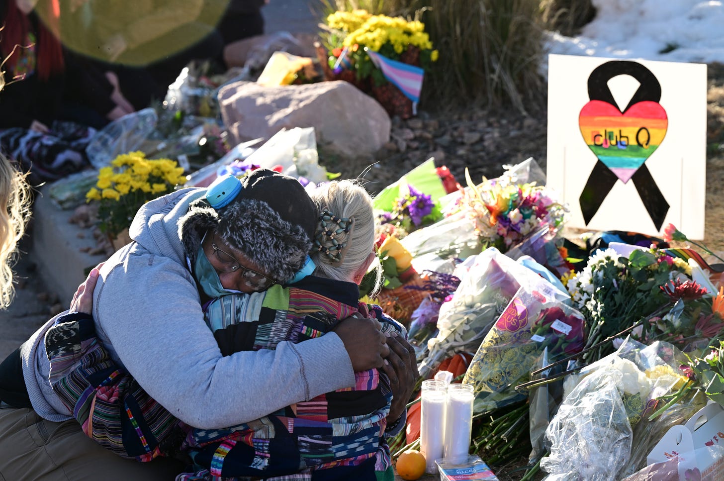 Two people embrace in front of a bed of bouquets of flowers, candles, and LGBTQ flags and signage.