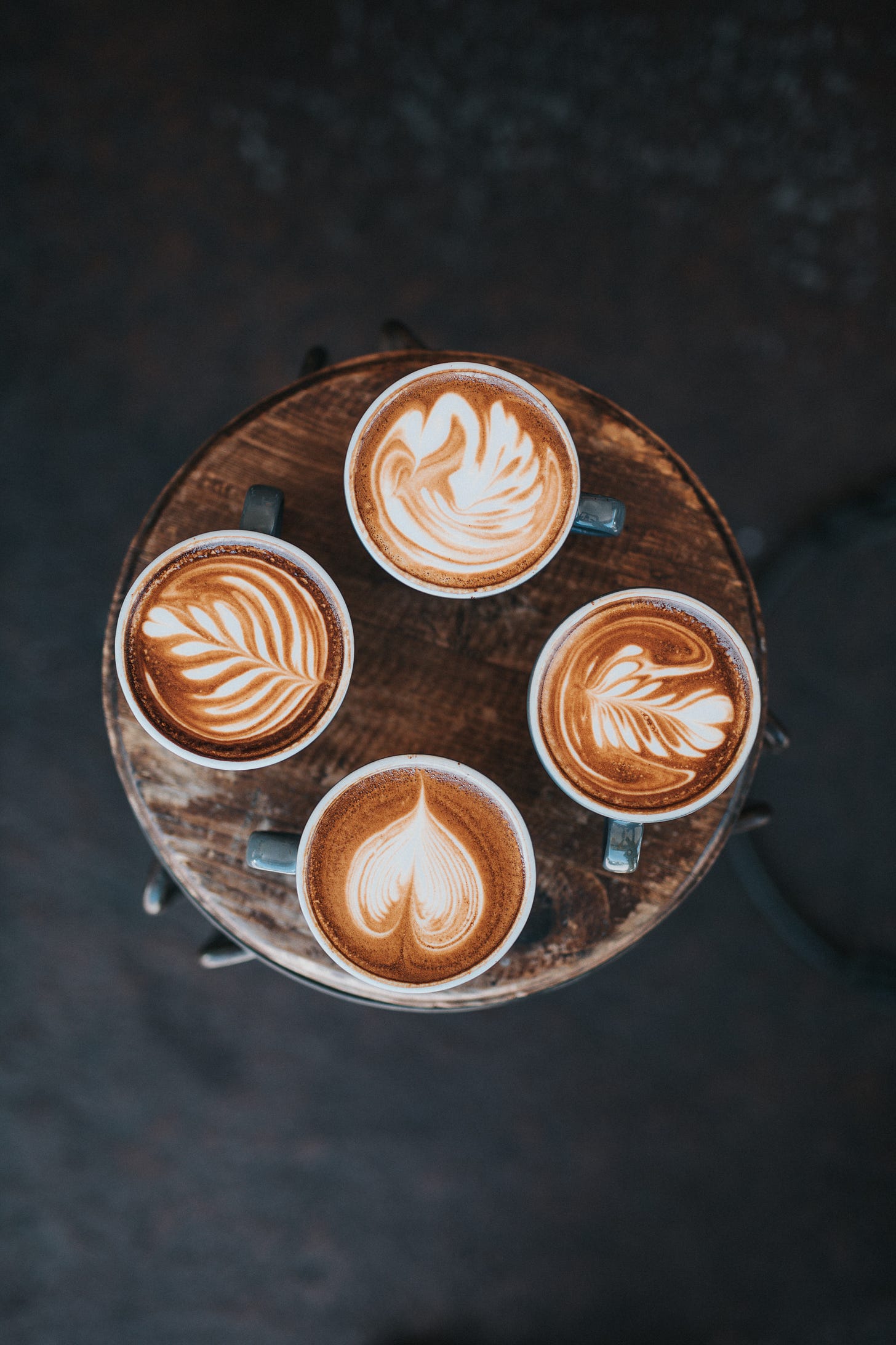 4 cups of coffee sit on a wooden table each with different foam art