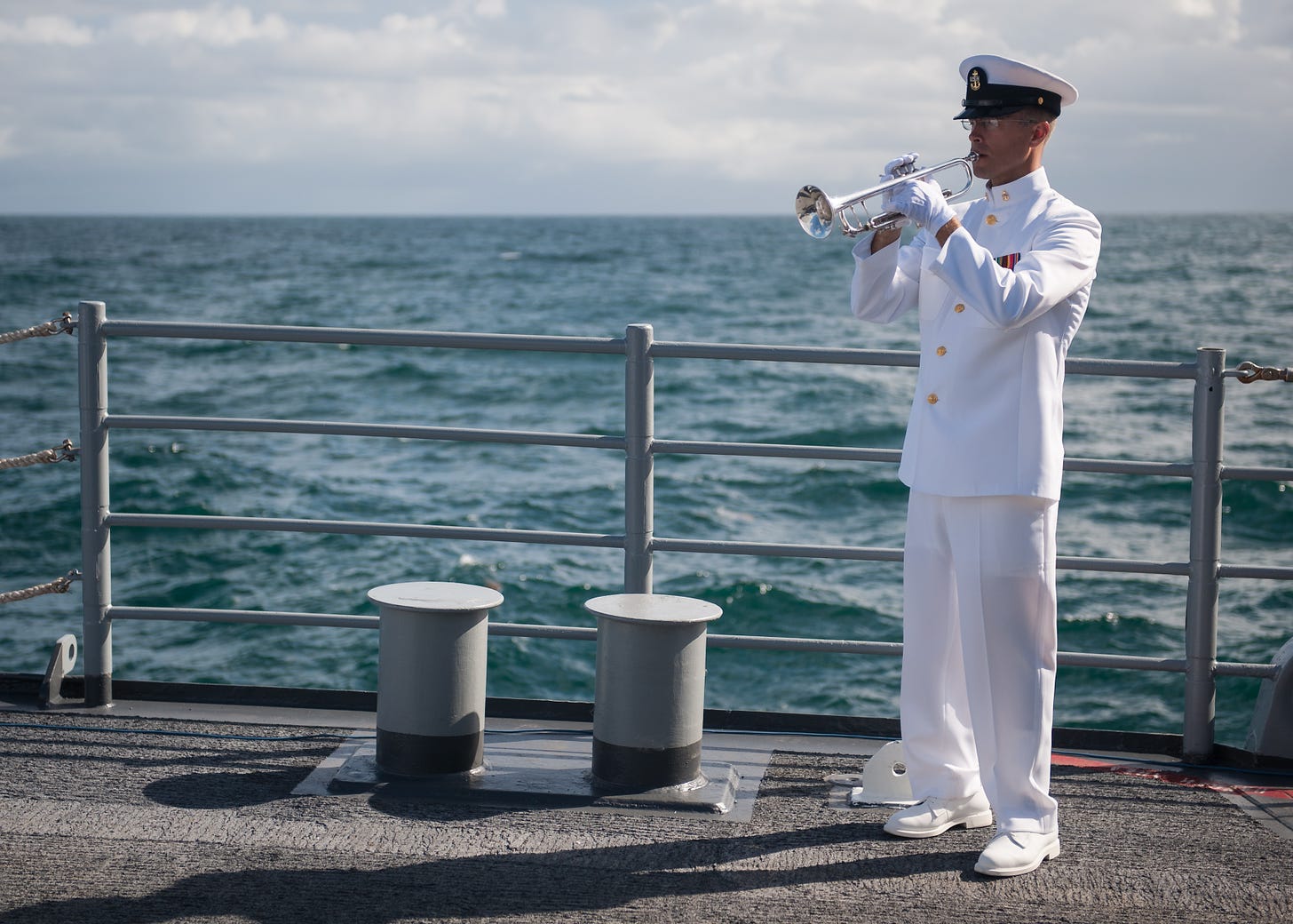 A man in a formal Navy uniform blows a trumpet on a ship with the ocean churning in the background.