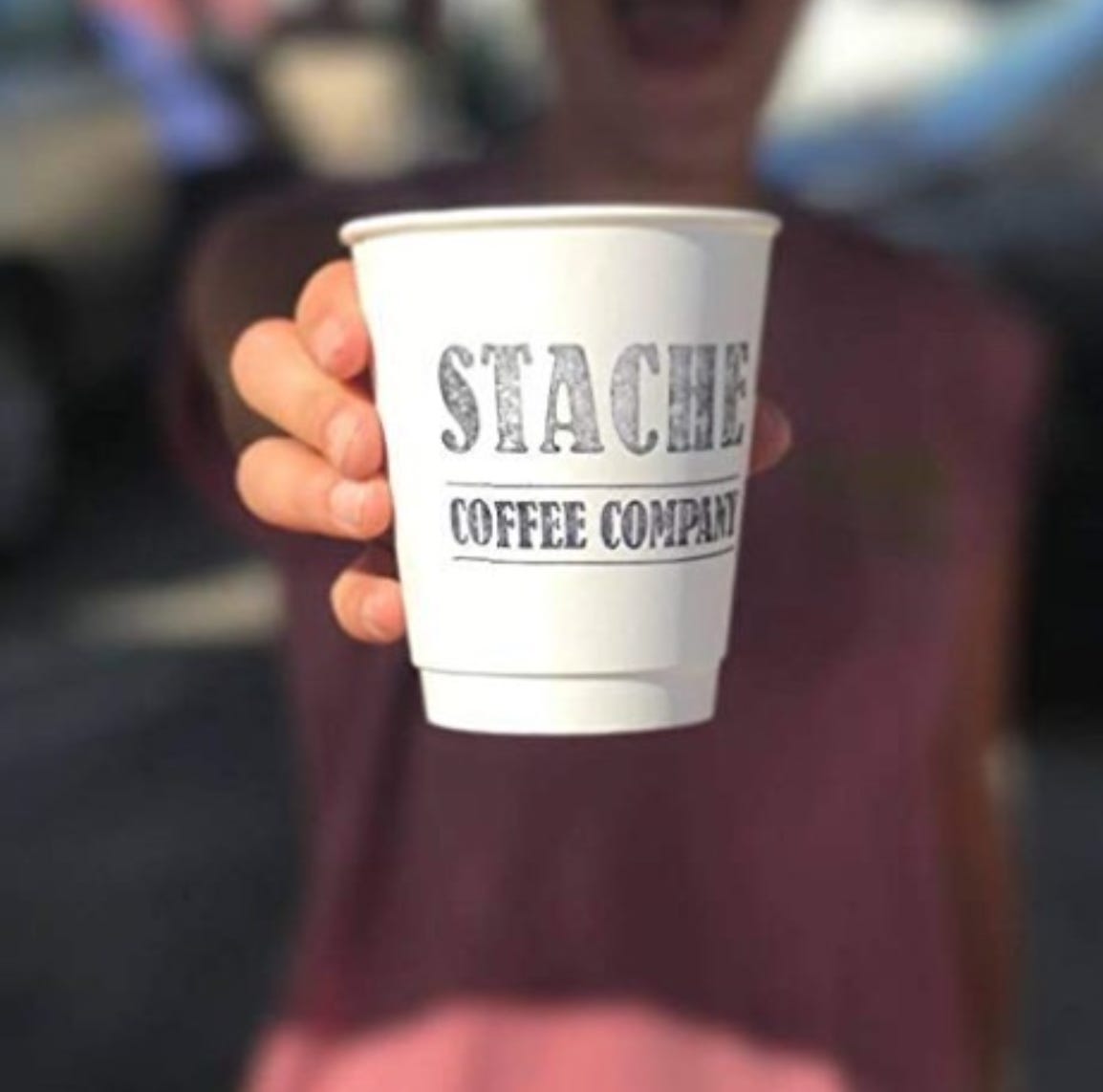 A white take out cup held up to the camera (background blurred) that has "Stache Coffee Company" inked on it.
