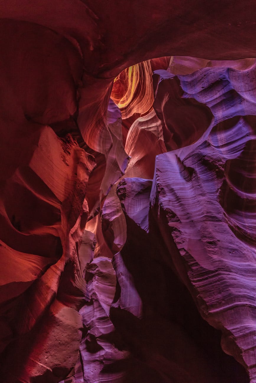 A carved rugged cave whose walls ae red and purple. Light enters it from above, The cave represents the carved heart of Jesus