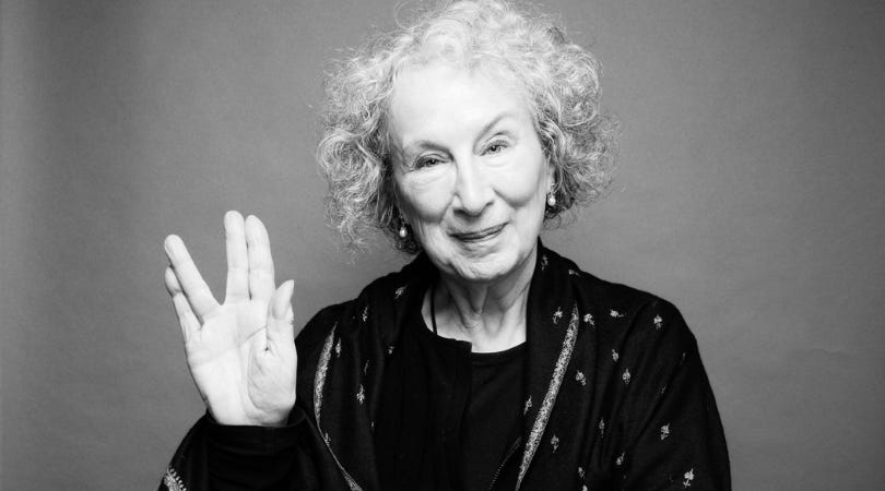 fotografia da autora Margaret Atwood em preto e branco. Uma senhora de cabelos bem brancos e cacheados, sorrindo e fazendo com a mão direita o sinal da saudação vulcana de star trek