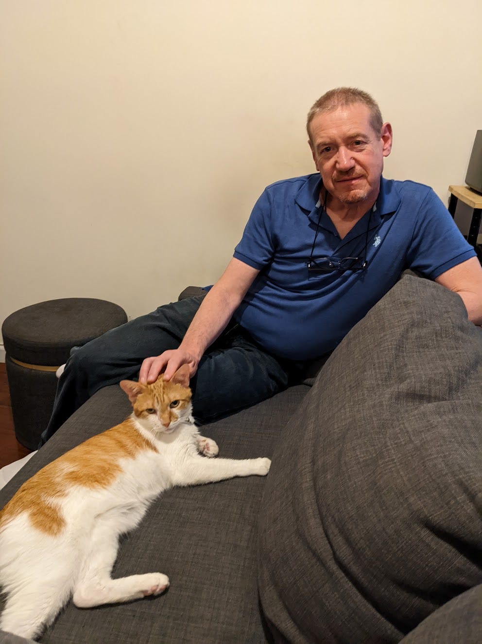 A white man with blond hair wearing a blue polo shirt and jeans sits on a couch petting a white and red cat