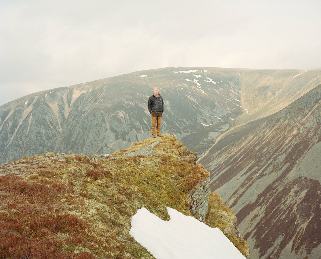 Thomas MacDonell stands atop a mossy peak, mountainous terrain rising behind him.
