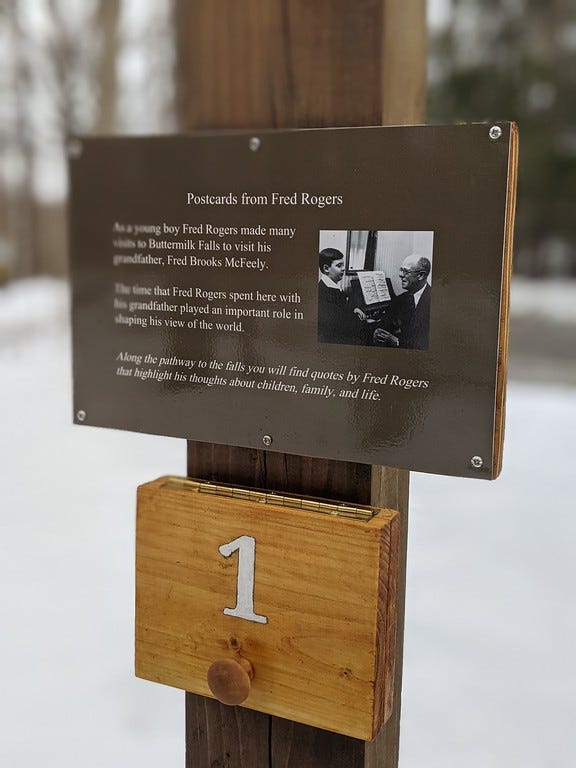 A photo of a wooden hiking trail sign attached to a wooden post featuring a photo of young Fred Rogers at the piano