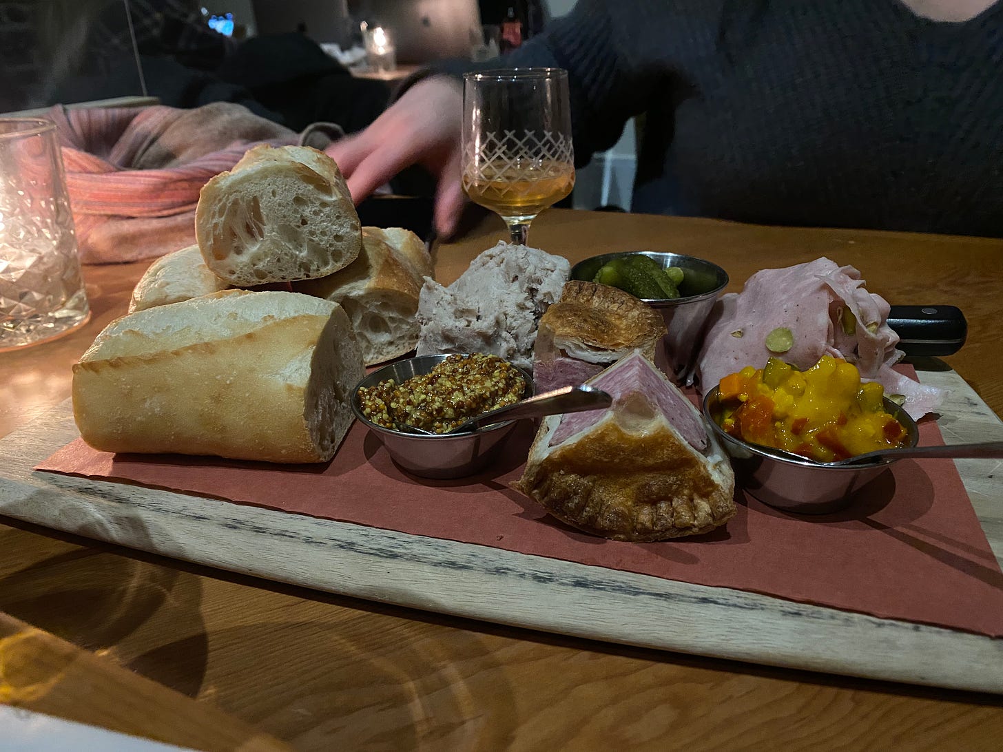 A wooden board of charcuterie: pieces of baguette, containers of mustard, pickles, and chutney, pieces of pork pie, and small piles of mortadella and duck rillettes. A candle sits to the left, and in the background is a small cocktail glass, nearly empty.