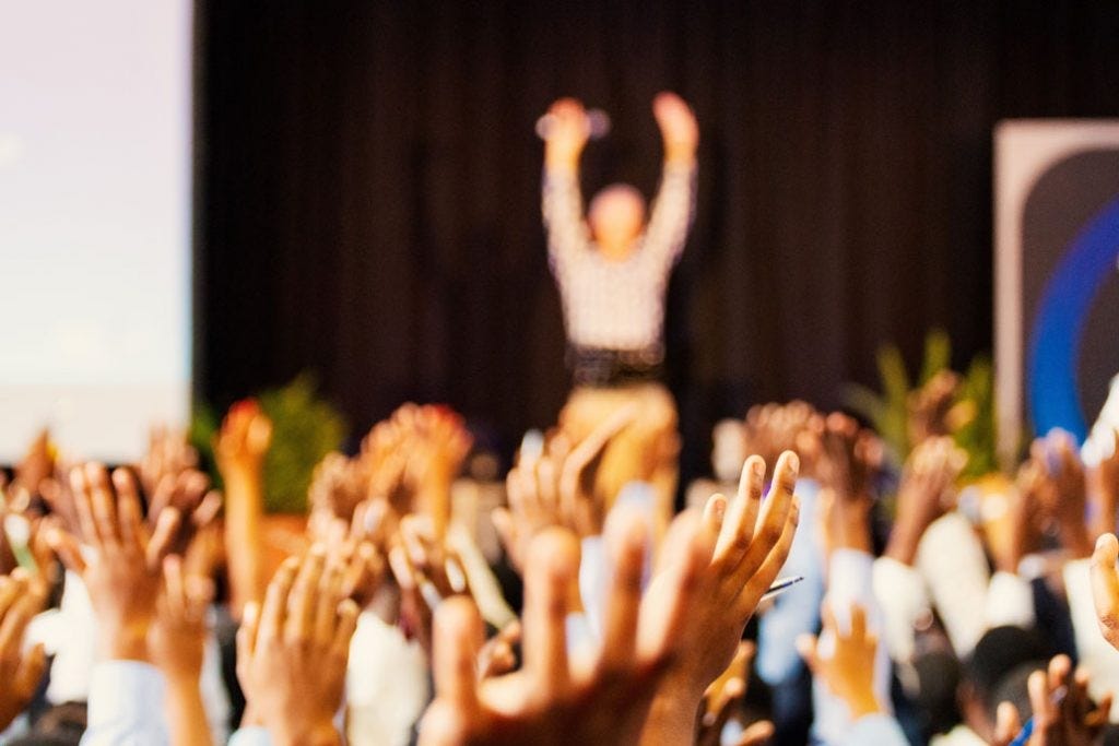 a crowd with their hands raised. in the background, there is a presenter on stage with their hands raised as well