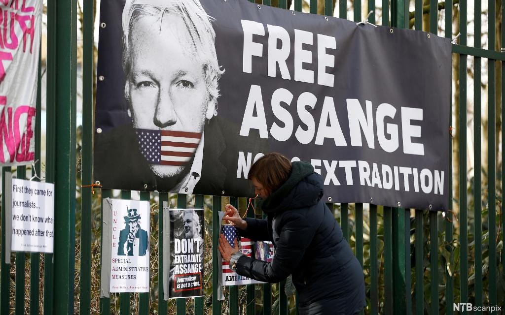En kvinne fester en stor plakat på gjerdet utenfor rettssalen The Old Bailey i London. Plakaten viser ansiktet til Julian Assange. Munnen hans er kneblet med en teip i fargene til det amerikanske flagget. Tittelen på plakaten er "Free Assange". Foto.