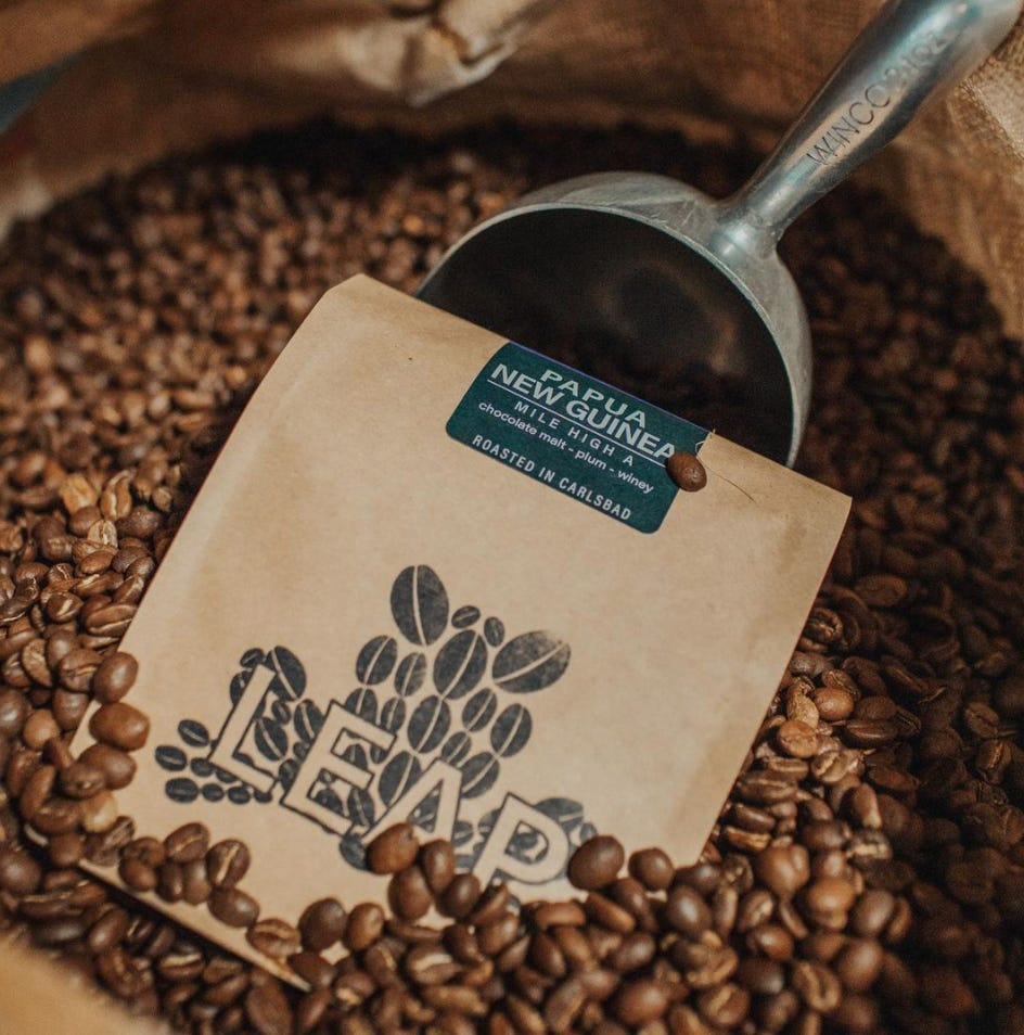 A brown bag of coffee beans with the Leap Coffee inked in the middle. The bag is half buried in a burlap sack of roasted coffee beans and propped up by a scoop.