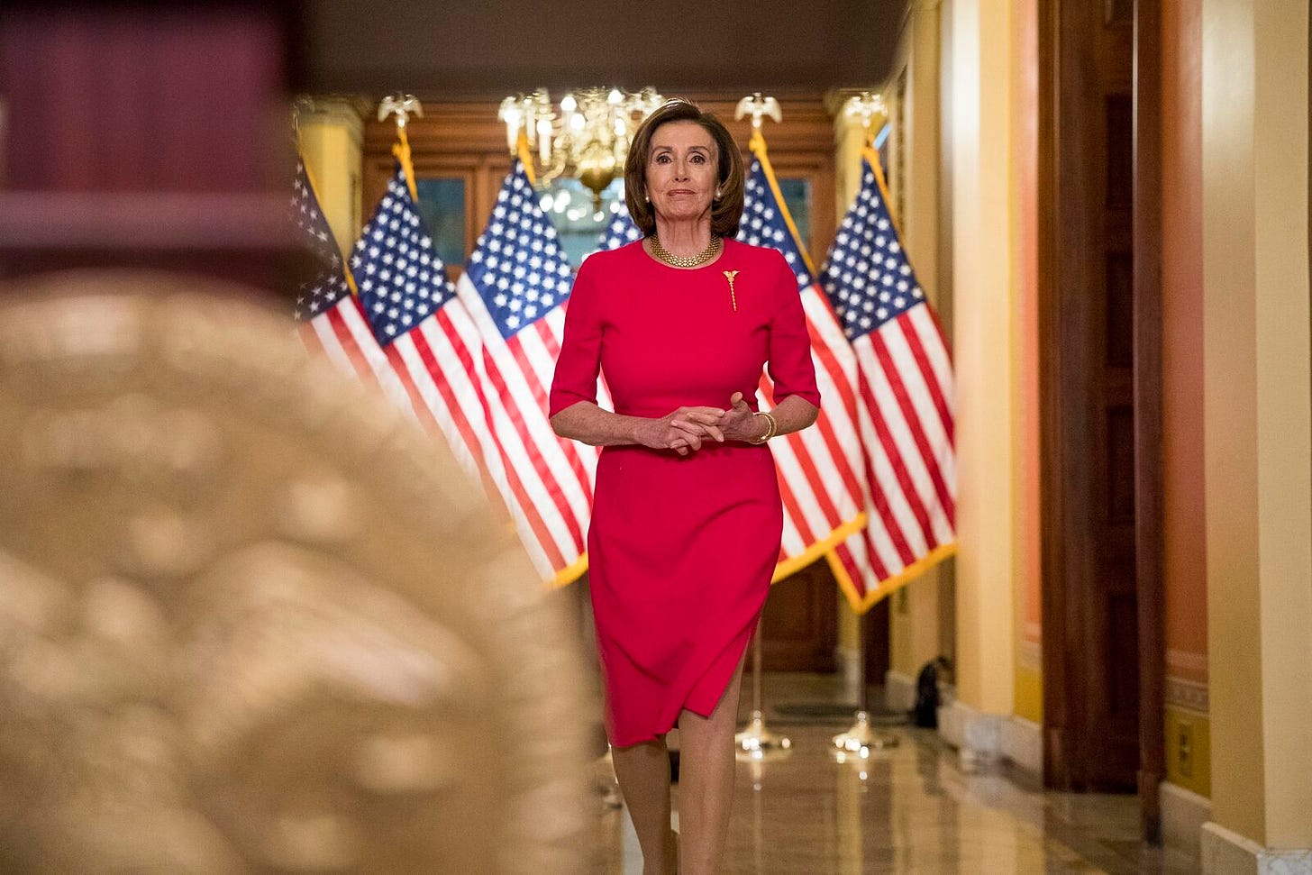 Nancy Pelosi, in a red dress, strides down a hallway in the US Capitol. Behind her, a half-dozen US flags fill the background.