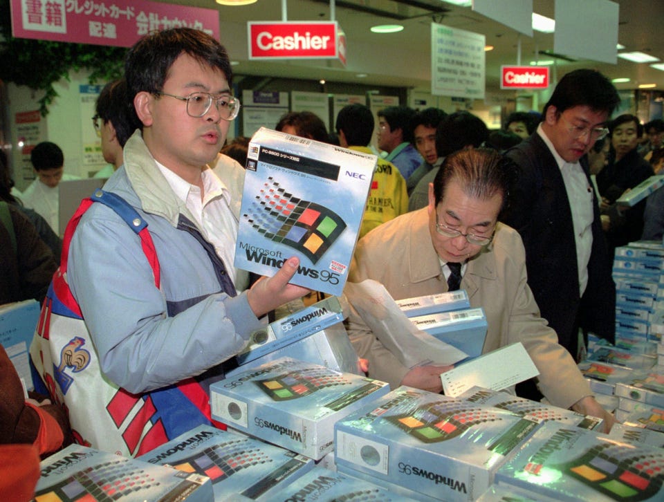Foto mostra uma pessoa segurando uma caixa com CDs ou disquetes de instalação do Windows 95, enquanto outras pessoas olham mais caixas do mesmo tipo em um stand de loja.