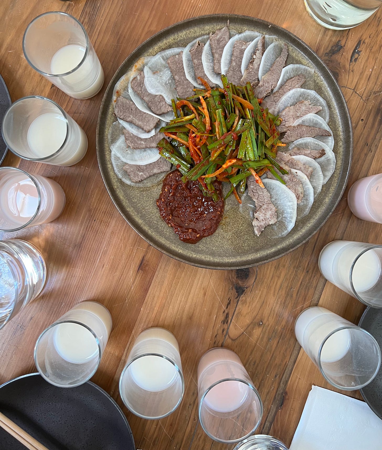 a picture of a table at a restaurant. on the table is a plate of sliced korean meat and radish. around the plate are nine small glasses filled with white and pink rice wine.