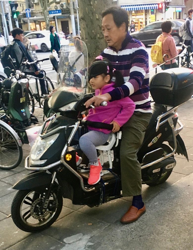 Dad & young child on a scooter on crowded sidewalk in Shanghai, neither with helmets.