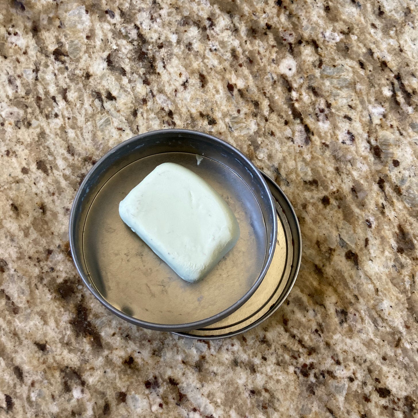 Visual description: A once-square light blue bar of solid deodorant with a rubbed-away rounded edge is held in a round, open metal container on a gray and brown granite countertop.
