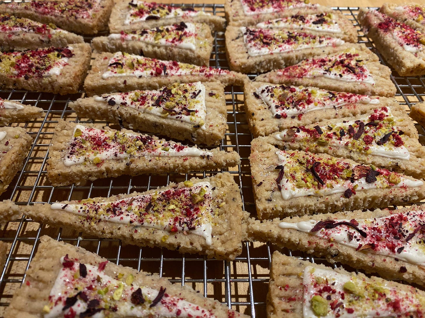 Two rows of triangle shaped shortbread cookies on a metal cooling rack. They are topped with white frosting, and a colorful mix of decorations: crumbled pistachio, dried hibiscus leaves, sparkling sugar, and freeze-dried raspberry powder.