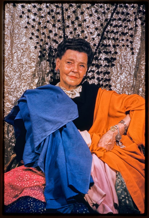 Portrait of Barbara Karinska in front of glitter curtain with blue, orange, and pink fabric draped over her. Her left hand is positioned on her hip.