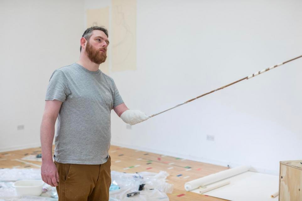 A white bearded person uses a very long paint brush as a prosthetic to paint a canvas just out of shot.