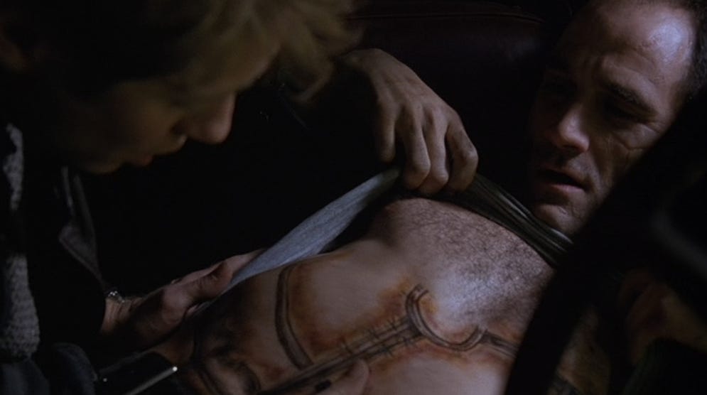 James (James Spader) examines the “prophetic” tattoo on Vaughan’s (Elias Koteas) abdomen. The tattoo is appropriately Cronenbergian in its ugliness. 