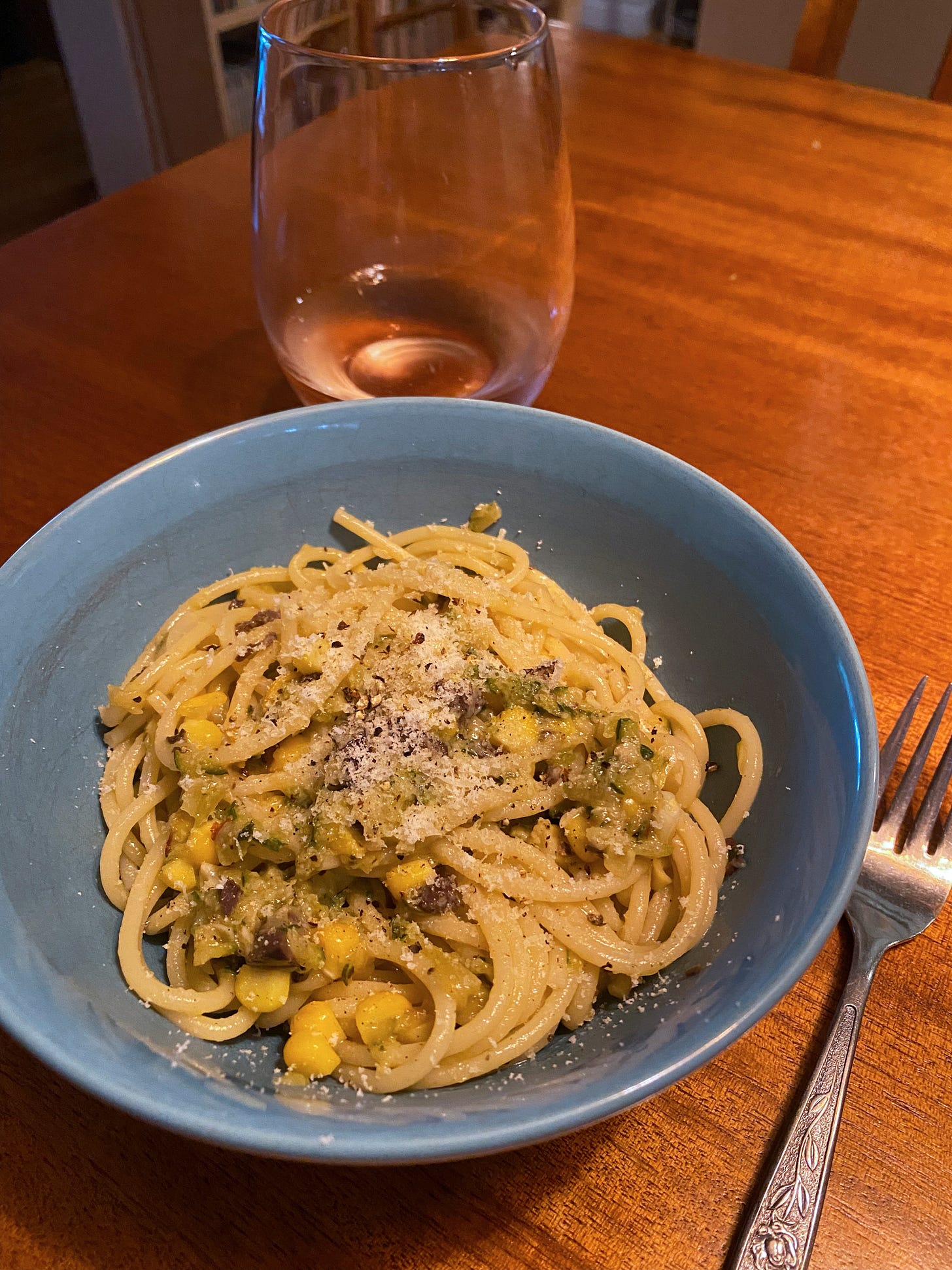 A blue bowl of spaghetti with zucchini, chopped olives, and corn. On top is a dusting of parmesan and cracked pepper. A stemless glass of rosé is on the table in the background.