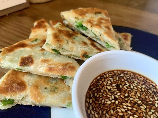 A pile of scallion pancakes next to a bowl of soy sauce with sesame seeds floating on it